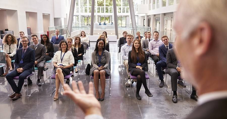 older man speaking in front of a diverse audience in a sunny white conference hall