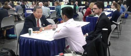 One man on one side of a table talking to two men on the other side of the table in a conference hall